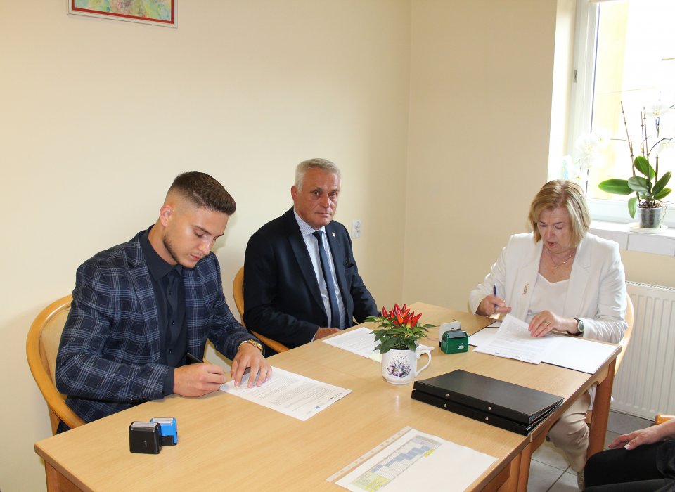 - umowa_szpital.jpg Dwóch mężczyzn siedzi na krzesłach, jeden z nich podpisuje dokument. Mężczyźni ubrani w garnitury. Obok nich siedzi kobieta z jasnymi włosami ubrana na biało, na stole leżą dokumenty, pieczątki oraz stoi niedużej wielkości kwiatek